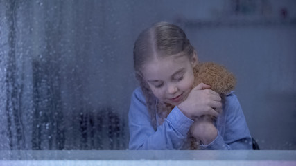 Fototapeta premium Lonely little girl playing with teddy bear at home in rainy weather, no friends