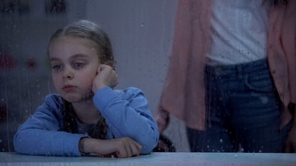Offended girl looking through rainy window, mum standing behind, punishment