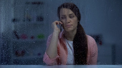 Upset middle-aged lady talking phone behind rainy window, bad news, conversation