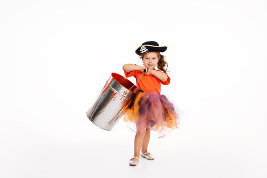 Happy child on Halloween. Funny little girl in carnival costumes of pirates standing with a big bucket of orange paint on white background. trick or treat