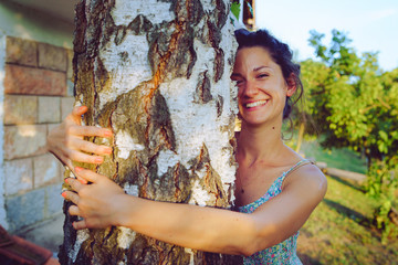 Young woman in summer dress hugging embracing tree trunk in her backyard in spring autumn fall day enjoying nature joyful happy smiling hug