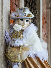 Lady in gold and white costume and masked face at Venice carnival. Highly decorative outfit with shiny details, eyes visible behind the mask. Old palace wall in background