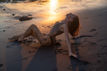 Beautiful sensual woman in sparkling swimsuit posing at the black sand beach during amazing golden sunset