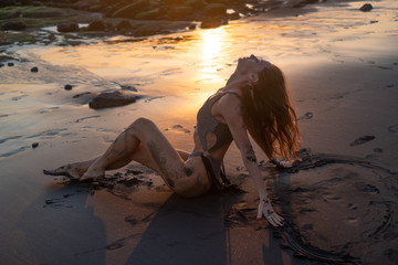Beautiful sensual woman in sparkling swimsuit posing at the black sand beach during amazing golden sunset