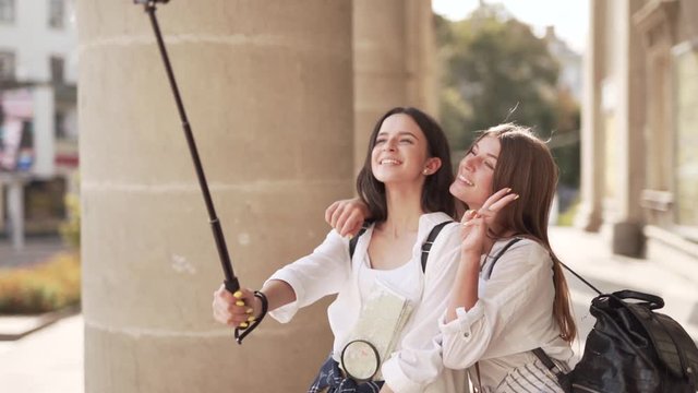 Two beautiful girls are standing on a stairs of some ancient building and making a selfie. They are tourists cause they are hoding a map of the city.