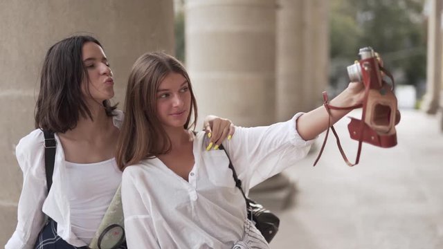 Two beautiful girls are standing on a stairs of some ancient building and making a selfie. They are tourists cause they are hoding a map of the city.