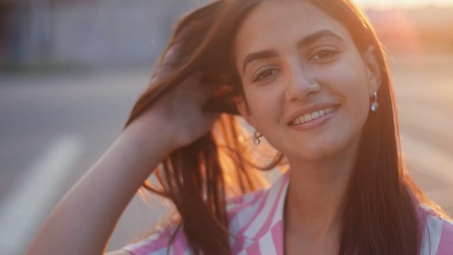 Beautiful young cheerful wooman smiling and playing with hair on the streer at sunset. Closeup portrait of positive girl outdoors in Slow motion