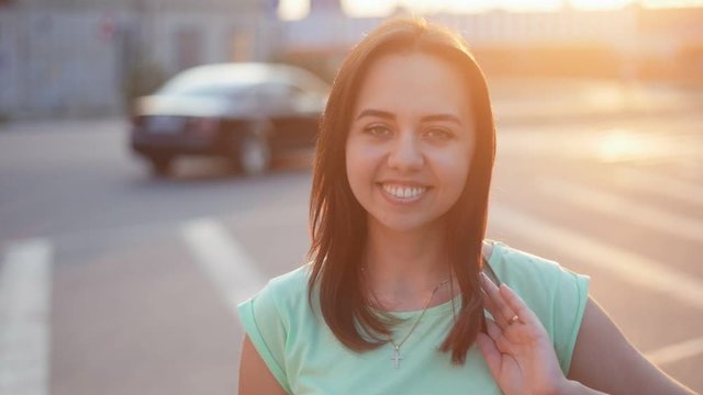 Beautiful young cheerful wooman smiling and playing with hair on the streer at sunset. Closeup portrait of positive girl outdoors in Slow motion