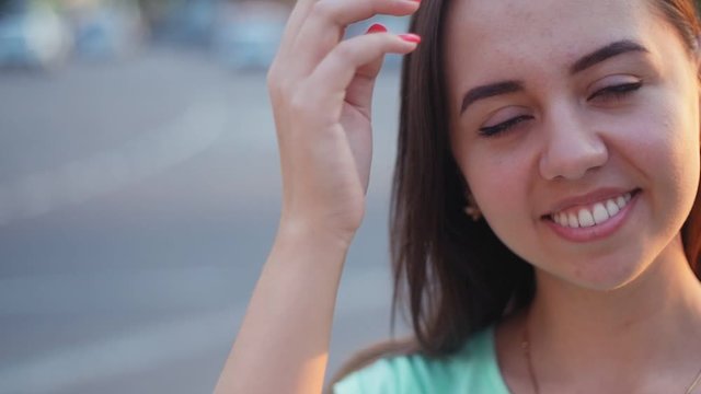 Beautiful young cheerful wooman smiling and playing with hair on the streer at sunset. Closeup portrait of positive girl outdoors in Slow motion