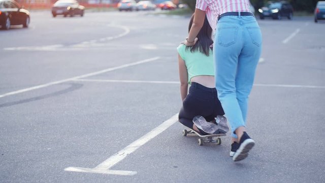 Positive girls having fun with scateboard on the parking at sunset in Slow motion. Two young cheerful women riding on the skateboard