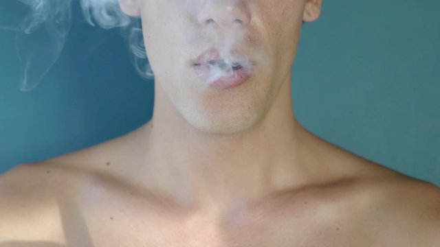 Young sportive man holding Juul (Vape) smokes an e-cigarette, breathes out streams of smoke in a close up view.