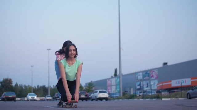 Positive girls having fun with scateboard on the parking at sunset in Slow motion. Two young cheerful women riding on the skateboard