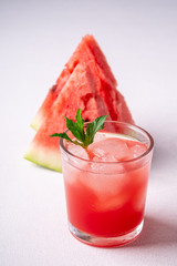 Fresh cold watermelon juice with ice cubes and green mint leaf in glass drink close to two slices of watermelon on white background, copy space, angle view, selective focus