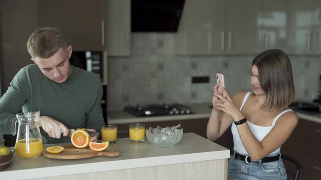 Young couple sitting at the kitchen counter cutting oranges to make juice while the girl takes a picture using her smartphone