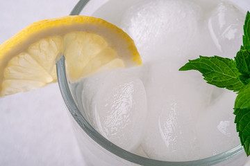 Fresh cold clear water drink in glass with ice cubes, lemon slice and mint leaf, on white gray background, macro, selective focus