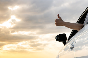 Man showing thumbs up/making Like / Ok sign with hand from car window with sunset sky, relaxing,...
