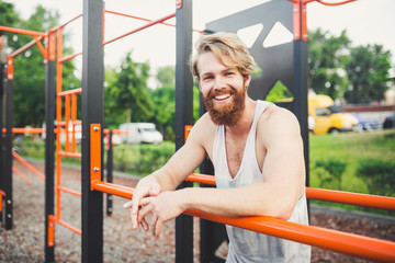 Fototapeta premium Portrait young bearded man standing on public sports ground and training on parallel bars. Handsome workout athlete. Portrait sporty man with beard, rest after training. Summer workout outdoors