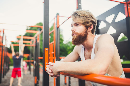 Portrait young bearded man standing on public sports ground and training on parallel bars. Handsome workout athlete. Portrait sporty man with beard, rest after training. Summer workout outdoors