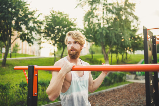 Portrait young bearded man standing on public sports ground and training on parallel bars. Handsome workout athlete. Portrait sporty man with beard, rest after training. Summer workout outdoors