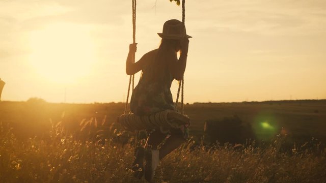 Happy child girl on swing at golden summer sunset. Silhouette of a young teenager girl swinging on the lone tree at nature, lifestyle. The concept of childhood dreams.