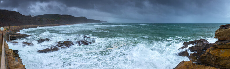 Fototapeta premium A huge ocean waves breaking on the coastal cliffs in at the cloudy stormy day. Breathtaking romantic panoramic seascape of ocean coastline. Peniche, Portugal.