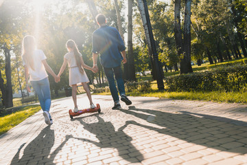 Rear view of young parents on sunny day walking in park with their cute little girl riding hoveboard