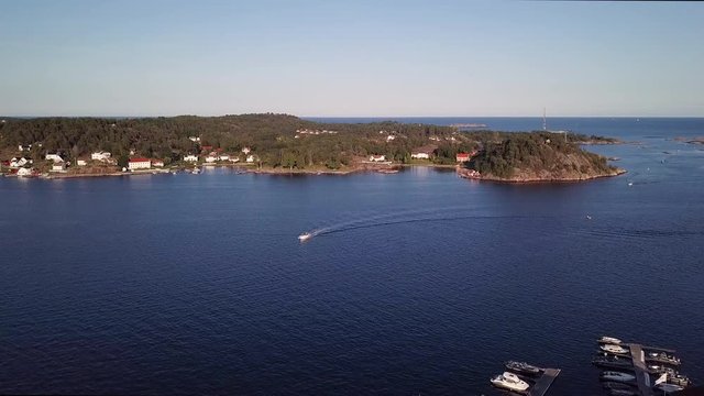 Aerial, descending, drone shot of a boat driving towards the harbor and buildings, in Ronnes village, near Grimstad city, on a sunny day, in Aust Agder, Norway