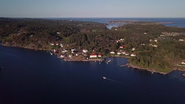 Aerial, drone shot, tilting towards a boat driving on the sea, in front of a small harbor and red buildings, in Ronnes, near Grimstad city, on a sunny day, in Aust Agder, Norway