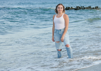 Stunning beautiful woman with blonde hair poses on beach in white tank top and blue jeans - laughing as wave hits