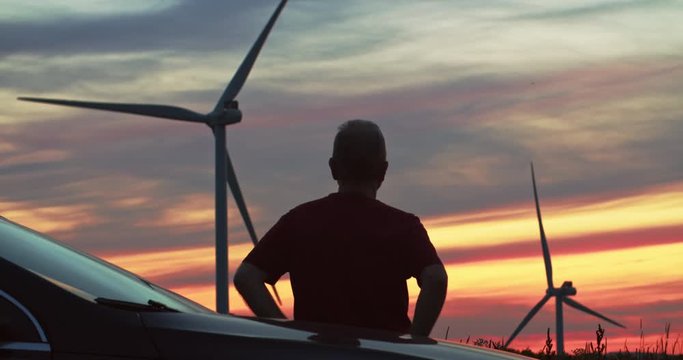 Back view man tourist observing rural landscape with eco-friendly windmills rotating in pink colorful skyscape.