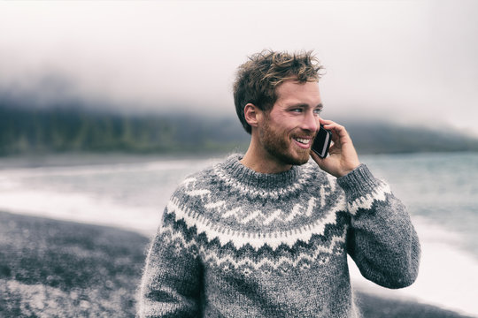 Phone man talking on smartphone in winter sweater walking on black sand beach in Iceland. Icelandic wool clothes. Technology mobile cellphone.