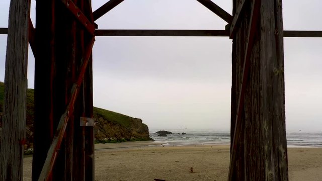 Aerial dolly back from the beach to Pudding Creek trestle basement, Fort Bragg, California. Drone shot on a cloudy day with sandy seashore near historical bridge.