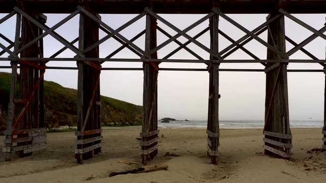 Aerial Dolly In  from the beach to Pudding Creek Trestle basement, Fort Bragg, California. Drone shot in a cloudy day with sandy seashore near historical bridge.