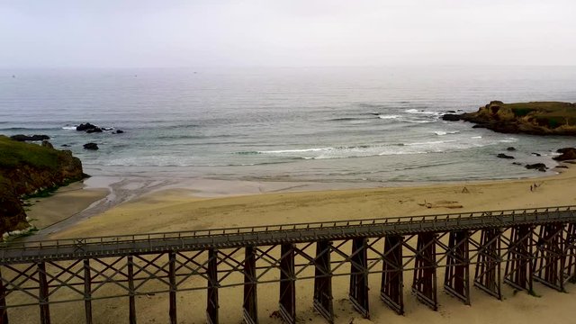 Drone Fly over Pudding Creek Trestle on sand beach. Aerial view from historical bridge to seashore with waves on a cloudy day.