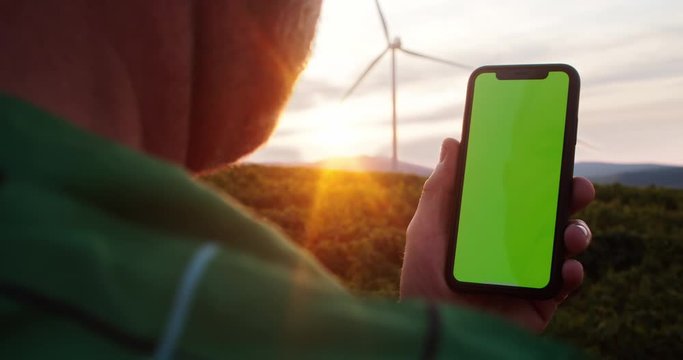 Close-up of farmer using mock up greenscreen smartphone outdoor. Busy man with mobile phone in rural field with eco-friendly wind turbines spinning and sunset sky.