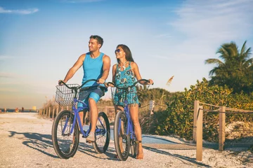 Foto auf Acrylglas Beach couple biking enjoying leisure sport recreational activity relaxing outdoors at sunset. Young woman and man riding bicycles on USA Florida vacation. Summer people lifestyle happy. © Maridav