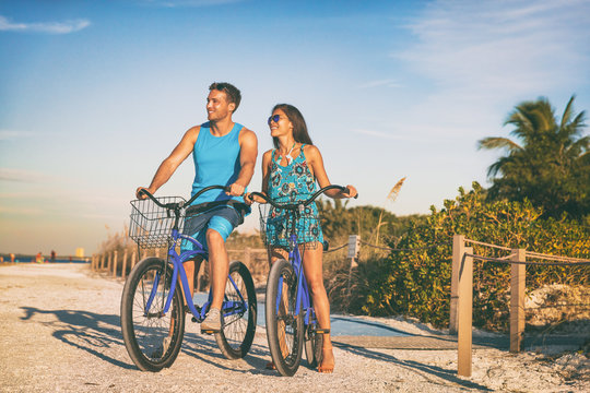 Beach couple biking enjoying leisure sport recreational activity relaxing outdoors at sunset. Young woman and man riding bicycles on USA Florida vacation. Summer people lifestyle happy.