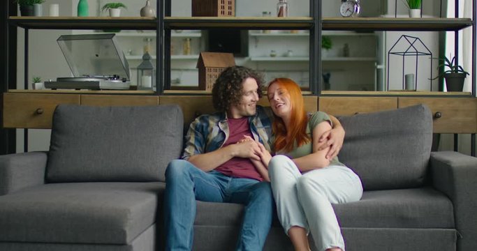 Young couple in love is sitting on sofa at home, in romantic mood, hugging, being happy, enjoying their relationships, man makes compliments to woman, Caucasian. 4K, shot on RED camera.