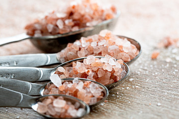 Close-up shot of coarse pink salt crystals on five steel measuring spoons, on the wooden background.Soft selective focus.