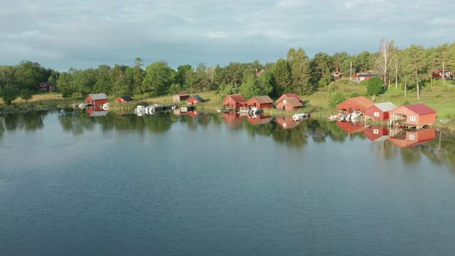Red boathouses along a shoreline. Beautiful huts and cabins. Camera pan along the coastline reveal the boat houses. Sunny summer morning.