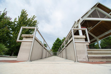 A low to the ground facing up angle of a concrete walkway surrounded by concrete walls and silver metal handrails leading up to a pavilion on a bright cloudy blue sky day.