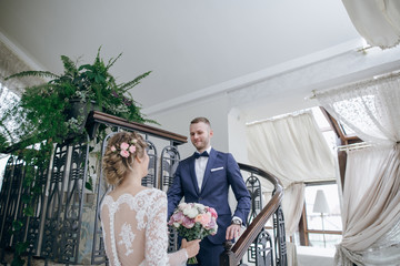 Fototapeta premium beautiful and elegant blonde bride in a long white dress with her handsome bride in a blue suit standing in the hotel room with bouquet of flowers