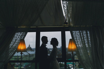 beautiful and elegant blonde bride in a long white dress with her handsome bride in a blue suit standing in the hotel room near window