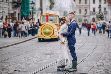 beautiful and elegant blonde bride in a long white dress with her handsome bride in a blue suit wallkig in a raining city with umbrella