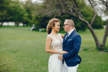 Fototapeta premium Beautiful bride in a long white dress. Handsome fiance in a blue suit. Couple in a summer park
