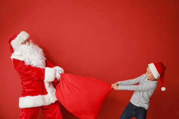 Little boy trying to take bag with gifts from Santa Claus against color background