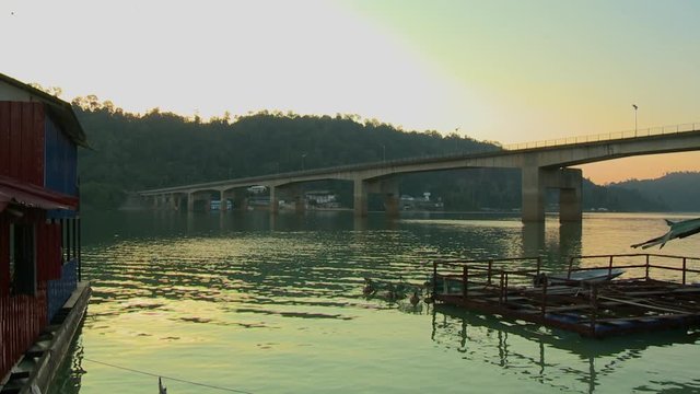 Wide low-angle evening still shot of a long concrete bridge crossing Temenggor Lake, and painted tin-roof jetty houses. At the bank are settlement houses, and silhouettes of a hilly forest above