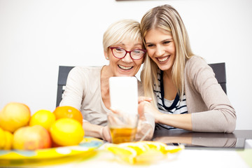 Senior mother and her daughter smiling and posing for a selfie while sitting by dinner table in bright room. Mother holding phone. Happy family moments at home.