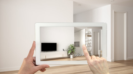 Augmented reality concept. Hand holding tablet with AR application used to simulate furniture and design products in empty interior with parquet floor, white living room with kitchen