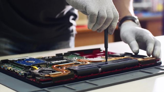Repairs and diagnostics breakdowns of laptop in professional service center. Electronic master detach accumulator of laptop in workshop, hands closeup. He unscrews battery from notebook screwdriver.